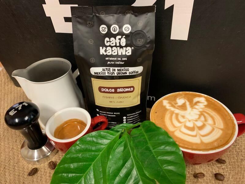 A bag of Café Kaawa Dolce Aroma, surrounded by a cup of espresso and a cappuccino with a pattern drawn on the foam, a coffee tamper, a milk jug, and decorated by green leafs and coffee beans