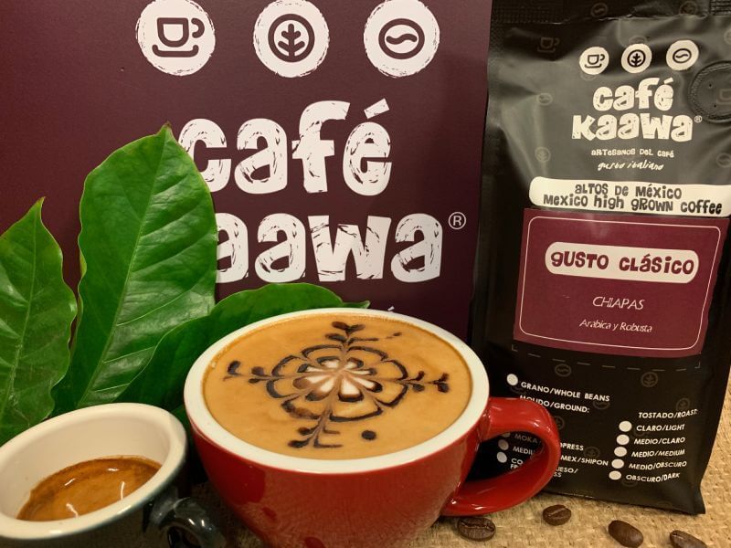 A bag of Café Kaawa Gusto Clasicó, a cup of espresso and a cappuccino with a flower design on the foam. Decorated by green leafs and coffee beans