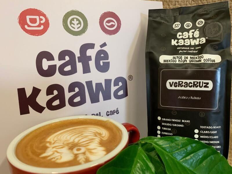Cup of coffee with intricate face design drawn on the foam, bag of Café Kaawa Veracruz and a sign with the Café Kaawa logo
