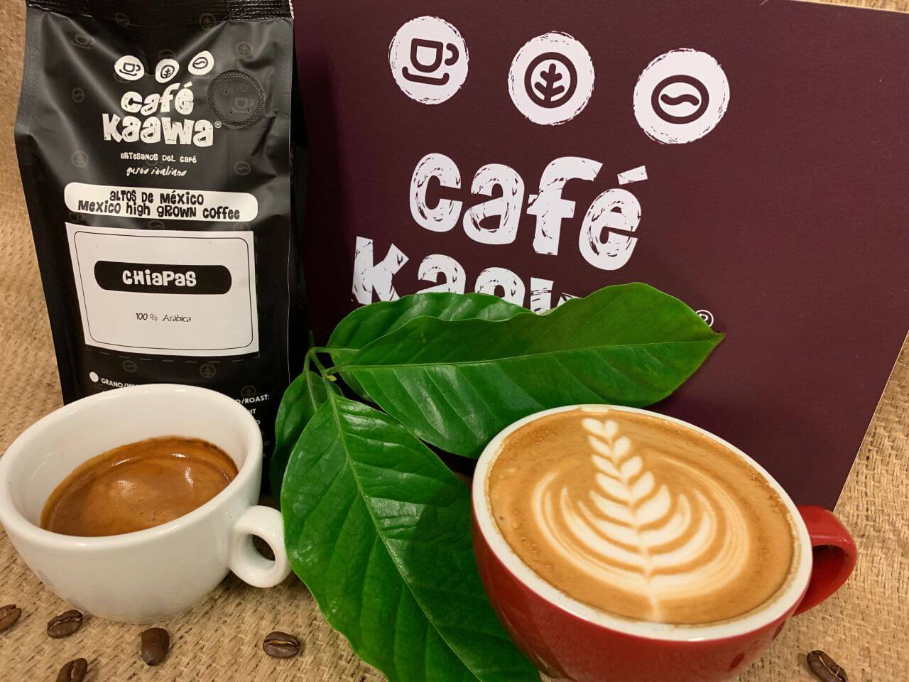 Two cups filled with coffee, one has a tulip pattern designed with foam milk. They're surrounded by green leafs, coffee beans, a Café Kaawa sign and a bag of Café Kaawa Chiapas