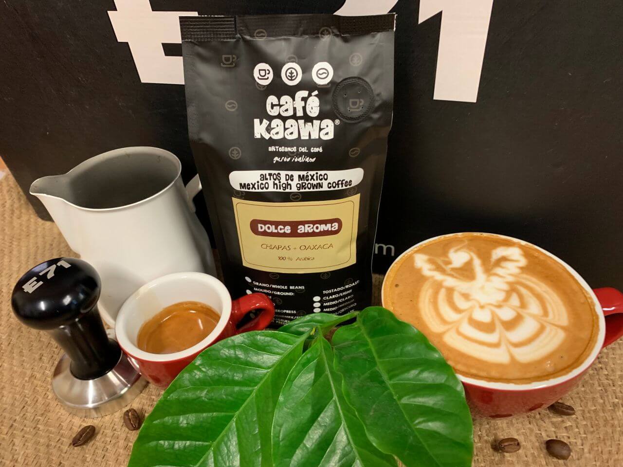 A bag of Café Kaawa Dolce Aroma, surrounded by a cup of espresso and a cappuccino with a pattern drawn on the foam, a coffee tamper, a milk jug, and decorated by green leafs and coffee beans