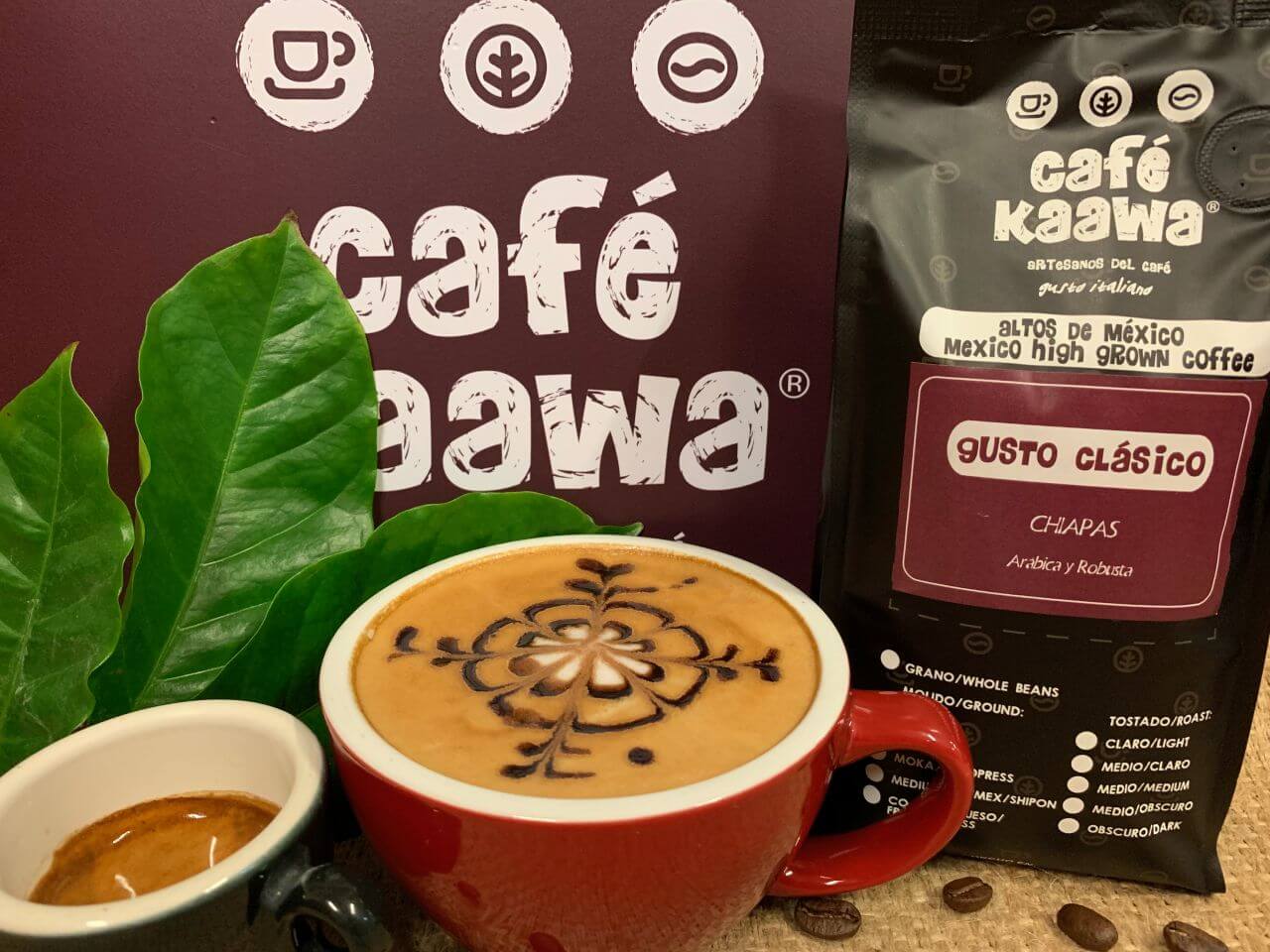 A bag of Café Kaawa Gusto Clasicó, a cup of espresso and a cappuccino with a flower design on the foam. Decorated by green leafs and coffee beans